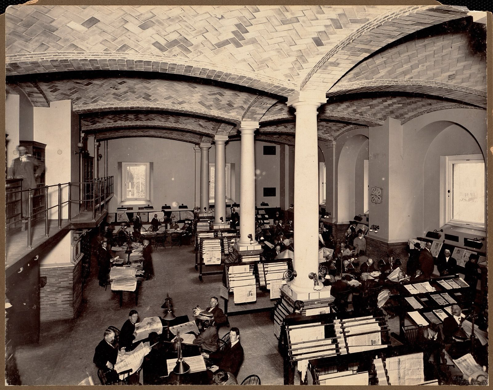 People reading newspapers at long racks and tables under a vaulted brick ceiling, Boston Public Library, around 1900