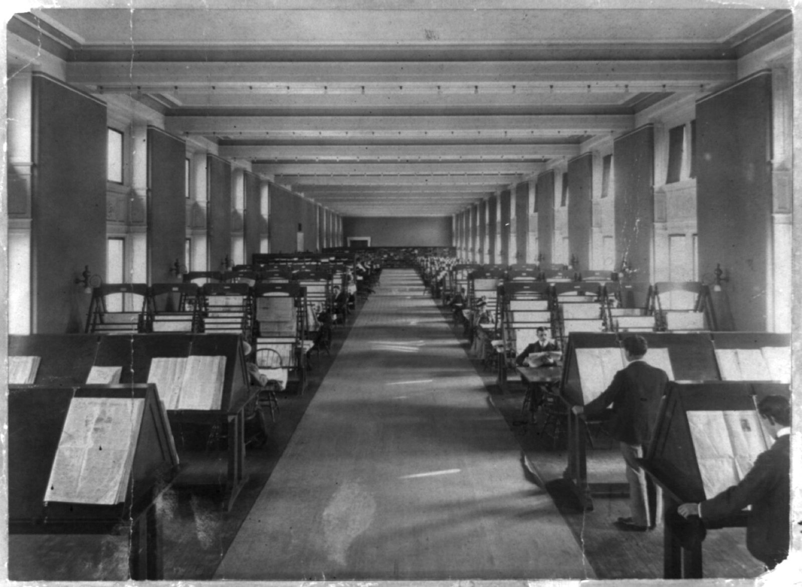 A long hall of slanted newspaper stands with readers bent over the papers, Library of Congress, around 1900